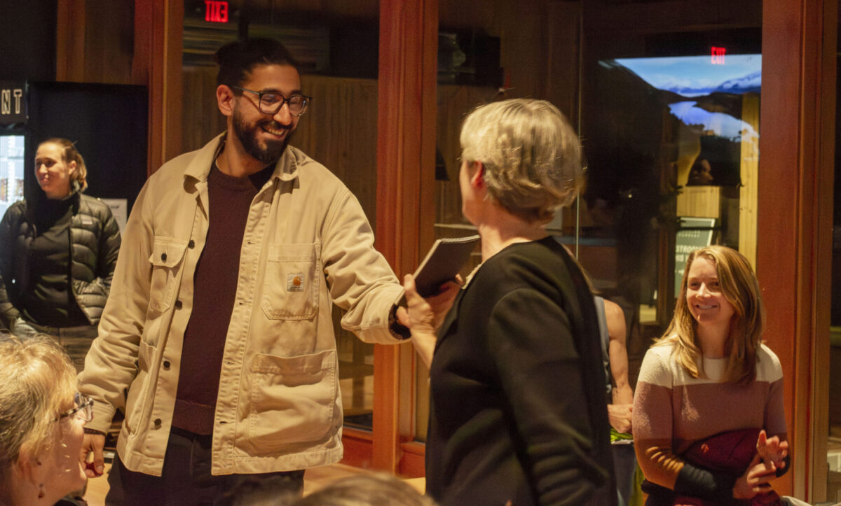 A smiling young man with a beard is handed a reporter's notebook at a community event.